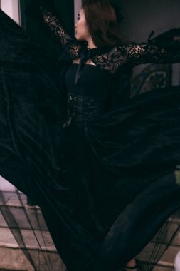 an Asian woman with a witch-like appearance flung her all-black skirt in the air as she stood in front of a cemetery before sunset