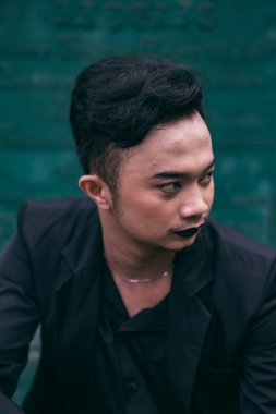 an Asian man in a black suit is sitting alone in front of the funeral monument with a very scary expression in the afternoon
