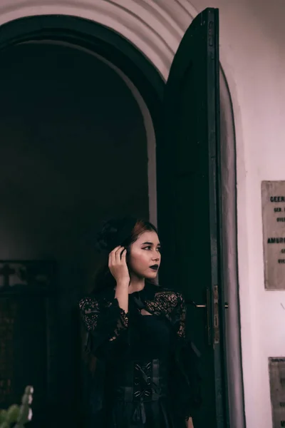 an Asian woman with lips and a black dress standing in front of an open door while visiting a cemetery in the afternoon