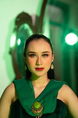 a beautiful Asian woman in a green dress has a very exotic face with ear and body jewelry while sitting in a green room