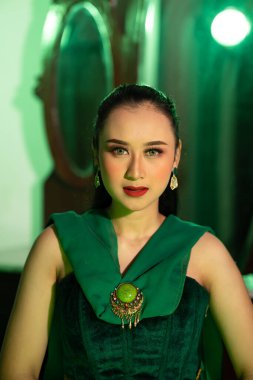 a beautiful Asian woman in a green dress has a very exotic face with ear and body jewelry while sitting in a green room