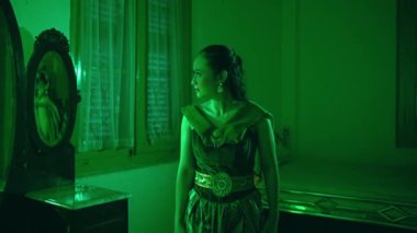 an Asian female dancer stands staring at a mirror with a sorry expression alone in her green room