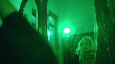 Müslüman bir kadın yeşil bir odada başörtüsü takarken aynada yüzüne bakar.