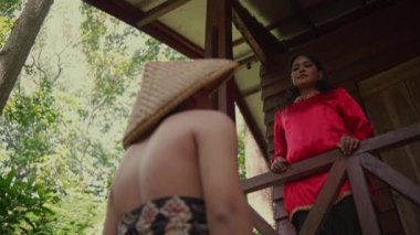Kırmızı tişörtlü Asyalı bir kadın, ormanın içindeki evin balkonunda birlikte vakit geçirirken hasır şapkalı bir adama bakıyor.