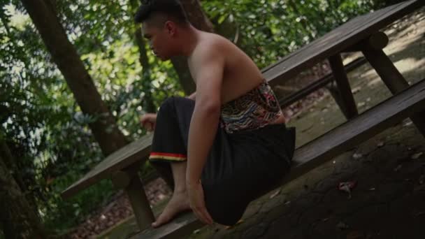 森の真ん中で動揺しながら黒い服を着た若い男が木のベンチに横たわっている — ストック動画