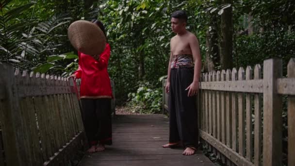 一位身穿红衣的妇女在森林中央的竹桥中央怒气冲冲地从她的伴侣手中扔出一顶竹帽 — 图库视频影像