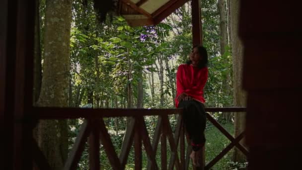 在丛林中的木屋前 身穿红色夹克的年轻女子坐在木制阳台的边缘上 — 图库视频影像