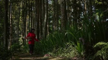 Güneşli bir günde, bahçenin yakınındaki ormanda yürüyen ve koşan Asyalı bir kadın. Kırmızı bir yağmurluk giyiyor..