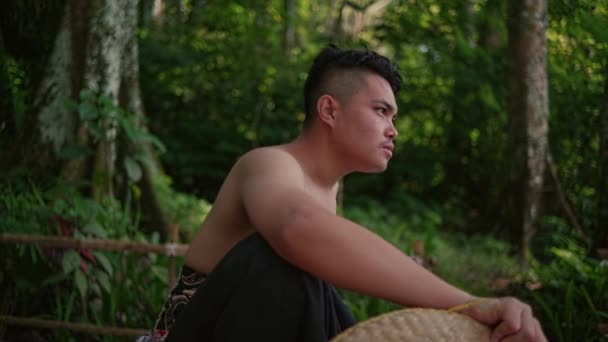 カメラを見てジャングルの中の木のテーブルとベンチに座っている若いアジア人の男性の肖像画 — ストック動画