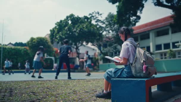 女子学生が学校の庭に座っていて朝バスケットボールをしている人たちを背景に — ストック動画