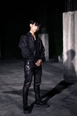 Siyah paraşüt ceketli Asyalı bir adam geceleyin bir binada çok erkeksi poz veriyor.