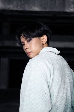 Siyah pantolonlu, şimşek desenli ve beyaz kazaklı Asyalı bir adam perili bir binada duruyor.