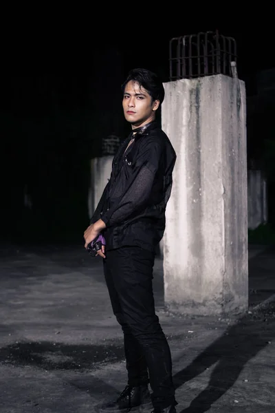 an Asian man dressed all in black and black hair posing as masculine in front of a building pillar