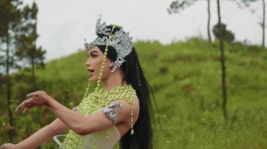 Göz kamaştırıcı bir fantezi kostümlü, gümüş bir taç giymiş karizmatik bir sanatçı dans ediyor ve kamerayla şakalaşıyor. Sahne, kültürel performansın, modern ifadenin ve efsanevi güzelliğin canlı bir karışımıdır..