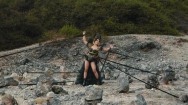 Dramatik siyah ve altın kostümlü güçlü bir cadı ya da kraliçe kollarını açmış, çorak, kayalık bir arazide taşlardan ve siyah kurdelelerden oluşan bir çember içinde büyülü bir ayin yapıyor..