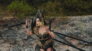 Zarif bir Karanlık Kraliçe 'nin yakın plan portresi. Süslü, tüylü başlığı ve dramatik makyajı görkemli zarafeti ve gizemli, güçlü bakışlarıyla poz verdiği yemyeşil bir bahçeye karşı sergileniyor..