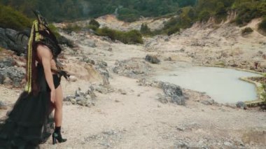 Dramatik siyah ve altın fantezi kostümlü bir kişi kayalık bir volkanik kraterin içindeki sütlü bir jeotermal gölün kenarında zarif bir şekilde poz verir. Sahne, yüksek modayla dünya dışı bir manzarayı harmanlıyor..