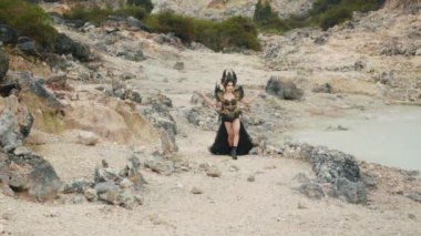 Geniş ve geniş açılı bir çekim, etkileyici siyah kostümlü güçlü bir kraliçeyi, yürürken ve kıraç, kayalık bir volkanik arazide zarifçe poz verirken takip eder. Sahne destansı, ıssız ve fantezi dolu..