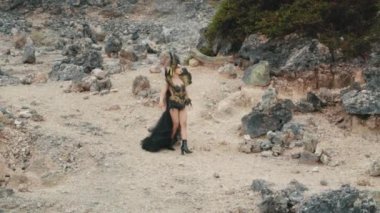 Yandan, zarif bir kraliçenin kayalık, volkanik bir arazide güvenle yürümesini izleyen bir izleme çekimi. Akan siyah elbise ve uzun sarı örgü dramatik görsel fanteziye ekleniyor..