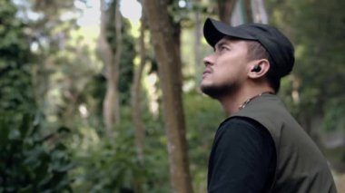 Siyah şapkalı Asyalı bir adam ormanın ortasındaki manzaranın tadını çıkarırken ahşap bir sandalyede oturuyor.