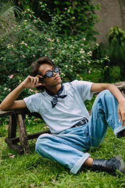 Kot pantolon ve güneş gözlüğü takmış Asyalı bir adam öğleden sonra yeşil çimlerin üzerinde oturmuş dinleniyor.
