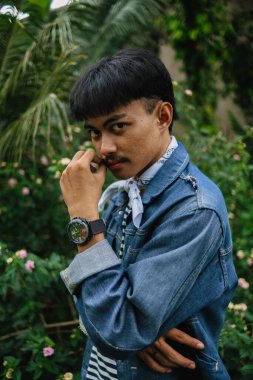 Kot ceketli ve çizgili tişörtlü Asyalı bir adam sabah çiçeklerle dolu bir bahçede düşünüyor.