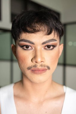 Asyalı bir adam beyaz gömlekli, kolları açık ve gece yüzünde makyaj var.