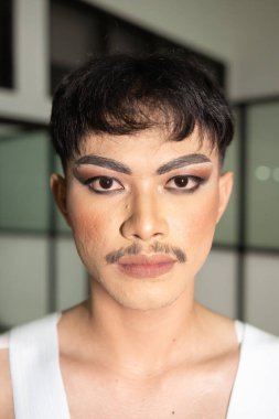 Asyalı bir adam beyaz gömlekli, kolları açık ve gece yüzünde makyaj var.