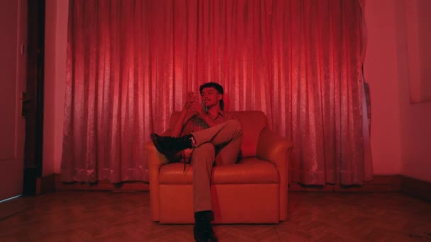 一位身穿复古服装的亚洲人孤零零地坐在夜晚鲜红的房间里的沙发上 — 图库视频影像