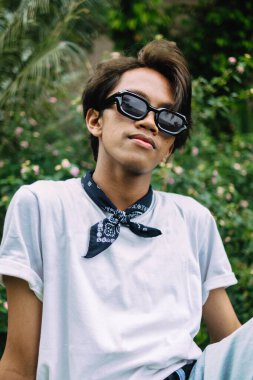 Güneş gözlüklü ve beyaz tişörtlü Asyalı bir adam sabah bir parktaki ahşap masada oturuyor.