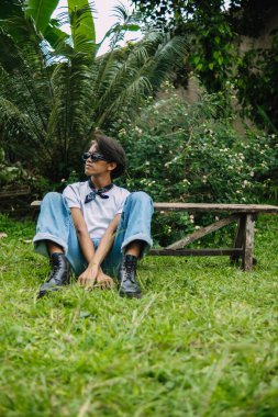 Kot pantolon ve güneş gözlüğü takmış Asyalı bir adam öğleden sonra yeşil çimlerin üzerinde oturmuş dinleniyor.