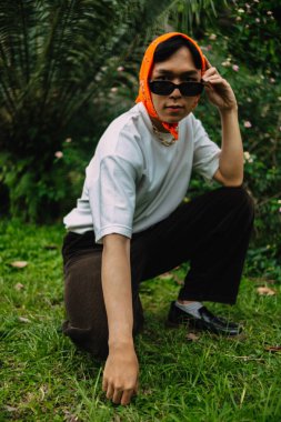 Genç bir çocuk güneş gözlüğü ve yeşil çimlerle dolu bir parkta turuncu bir bandana takıyor.
