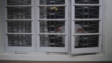 Beyaz tişörtlü ve güneş gözlüklü bir adamın beyaz, retro tarzı bir pencereyi açtığı orta boy bir fotoğraf. Dekoratif güvenlik parmaklıklarına yaslanır ve dışarıya bakar, gözlemci, düşünceli ya da gizemli görünür..