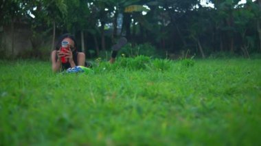 Güneş gözlüklü bir gencin yeşil çimlere uzanmış kırmızı bir tenekeden patates cipsi yerken geniş açılı görüntüsü. Güneşli bir günde dışarıda dinleniyorlar. Öndeki çimenler bulanık.