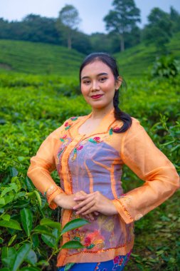 Turuncu elbiseli bir çay yaprağı çiftçisi sabah çok güzel bir çay bahçesinin önünde poz veriyor.