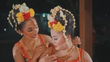Bir kadın oturmuş Endonezyalı bir dansçının arkasında altın kolyesinin ayarlanmasına yardım ediyor. Her ikisi de gülümsüyor, geleneksel kostümleriyle Java dansı gösterisi için hazırlanıyorlar..