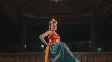 Canlı geleneksel bir Java dans kostümü giymiş güzel bir Asyalı kadına yavaşça zum yap. Gece bir çadırın ahşap sahnesinde zarif bir şekilde poz ver ve kameraya güven içinde bak..