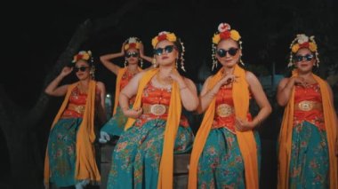 Bir grup geleneksel Asyalı dansçı, geceleri turuncu kostümlü ve güneş gözlüklü arkadaşlarıyla oturuyorlar.