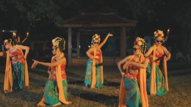 Geleneksel Endonezya kostümleri, çiçek başlıkları ve güneş gözlükleri takan beş kadın dansçı gece vakti açık havada senkronize bir dans gösterisi yapıyorlar..