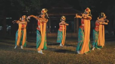 Geleneksel kostüm ve güneş gözlüğü takan beş Endonezyalı kadın dansçıdan oluşan bir grup, gece çimenli bir çimenlikte, arkalarında bir pavyon ve meşalelerle senkronize bir kültürel dans sergiliyorlar..