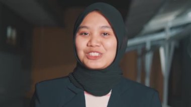 Genç bir Güneydoğu Asyalı profesyonelin tesettür ve spor ceketli pozu. Doğrudan kameraya konuşurken neşeli bir şekilde gülümsüyor. Vloglar ya da şirket videoları için ideal..