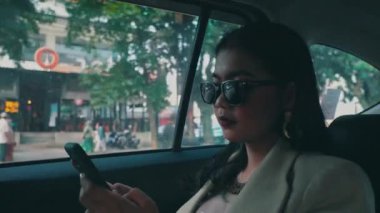Güneş gözlüğü takan şık bir iş kadını akıllı telefonunu kullanır. Sonra da şehirde bir arabanın arka koltuğunda giderken telefona cevap verir..