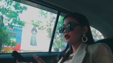 Ciddi, zarif giyimli, güneş gözlüğü takan bir kadın, bir arabanın arka koltuğunda giderken akıllı telefonuyla önemli bir telefon görüşmesi yapar..