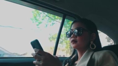 Güneş gözlüğü takan zarif bir iş kadını telefonunu kullanıyor ve bir arabanın arkasında giderken önemli bir çağrıya cevap veriyor..