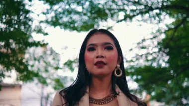 Düşük açı, kendine güvenen, zarif giyimli, ağaçların altında poz veren Asyalı bir kadına yakınlaşıyor. Saçlarına dokunur ve etrafına dik dik bakar..