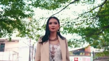 Yeşil ağaçların altında makyajlı şık bir Asyalı kadına zum yap. Saçına dokunuyor ve dikkatlice etrafına bakıyor. Düşük açılı güzellik portresi.