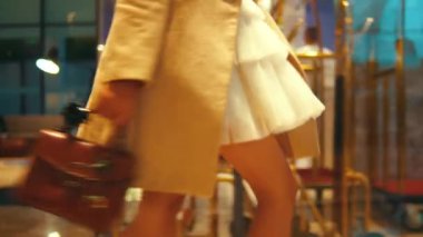 Gece vakti otelin lobisinde yürürken bej ceketli ve beyaz elbiseli bir kadının çantasını takip ediyormuş. Harekete ve tarzına odaklan.