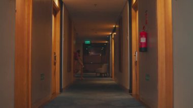 Statik geniş açı modern bir otel koridorunu gösteriyor. Bej ceketli bir kadın odadan çıkar, koridoru geçer ve en uçta durur..