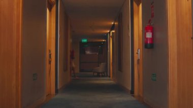 Bir otel ziyaretçisi geceleri oda arayan sarı ışıklarla dolu bir koridorda yürüyor.