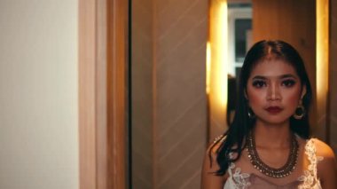 Göz kamaştırıcı beyaz elbiseli ve kalın makyajlı güzel bir kadın doğrudan kameraya bakar, sonra ahşap kapıyı kapatarak yüzünü saklar, bir yarısını görünür halde bırakır..